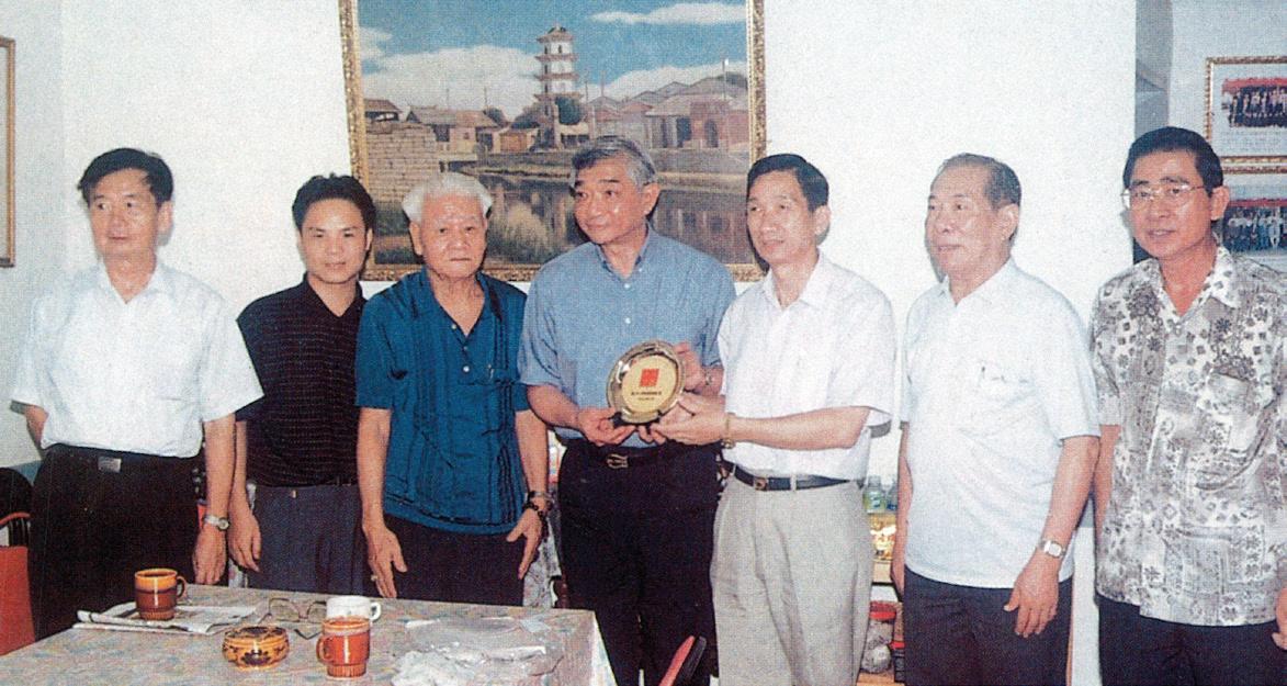 1998-年8-月，公会接待晋江市副市长吴松茂及侨办副主任吴晋居。图为公会副主席高泉乐接受吴副市长赠送的纪念盘。左起：颜呈註、颜期巢、吴松茂、高泉乐、颜期伟、吴晋居、陈方圆。